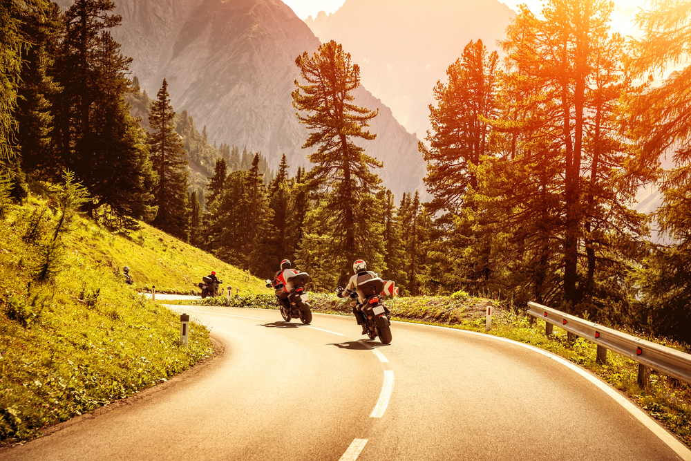 Group of motorcyclists riding on curves mountainous road, race of motorbike in Alps, beautiful pine forest, red sunset light, extreme sport concept